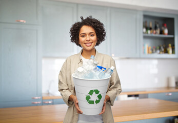 recycling, waste sorting and sustainability concept - smiling woman holding recycle bin with plastic bottles over home kitchen background - Powered by Adobe