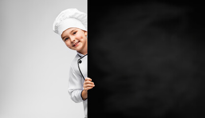 cooking, culinary and profession concept - happy smiling little boy in chef's toque and jacket with black chalkboard over grey background
