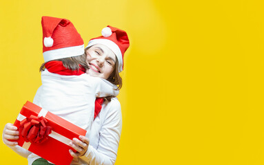Fototapeta premium Happy Family of Two Positive Caucasian Girls Mother and Daughter In Festive Santa Hats Having Fun While Embracing And Holding Wrapped Giftbox Behind On Yellow Background.