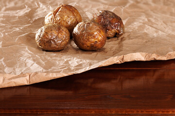 Whole baked potato tubers on brown kraft paper on a dark wooden table. Angle view. Free space for text.