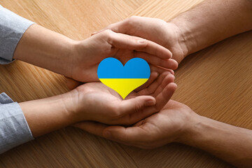 Stop war in Ukraine. Man and woman holding heart shaped symbol with colors of Ukrainian flag in hands at wooden table, closeup