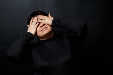 a brunette woman in black clothes covered her face with her hands on a black background