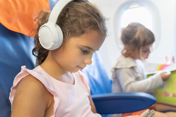 Kid schoolgirl in headphones watches cartoons in a phone on an airplane. little girl travels in an...