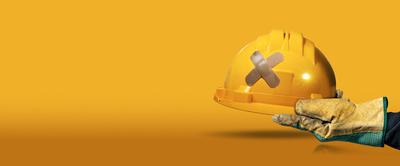 Manual worker with protective work glove holding a yellow safety helmet with a cross bandaid...