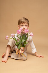 a charming blond four-year-old boy in beige overalls and a white shirt sits on a beige background