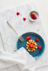 Vibrant blue plate with waffle with strawberries and raspberries on marble table and white background