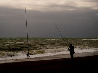 A silhouetted sea angler, with rods in the sand, fishing from a cold sandy beach at Fuengirola, southern Spain. Stormy waves breaking under a dark cloudy sky. 