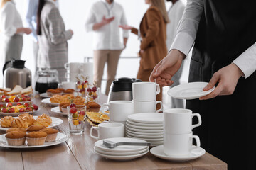 Waitress near table with dishware and different delicious snacks during coffee break, closeup