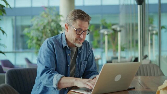 Happy middle age businessman using laptop computer in office. Older casual man working online on terrace of cafe, smiling.