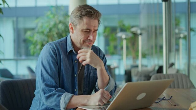 Happy middle age businessman using laptop computer in office. Older casual man working online on terrace of cafe, smiling.