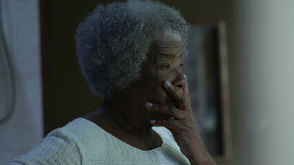 A preoccupied senior black woman standing by window looking out a worried African older lady