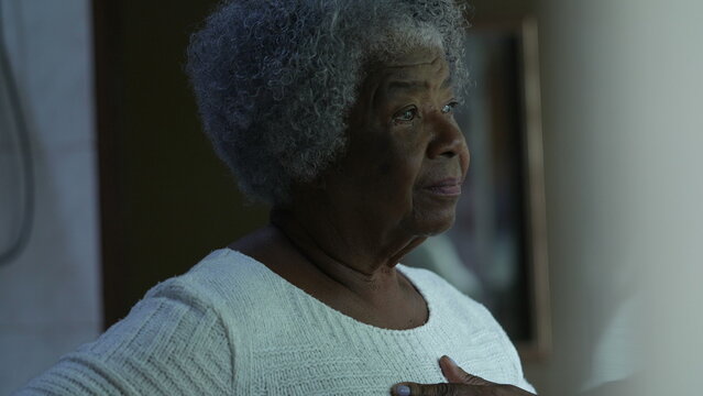 A preoccupied senior black woman standing by window looking out a worried African older lady