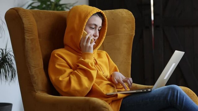 Stylish teenager girl sits in armchair with laptop computer and talking by mobile phone