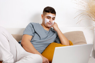 Young man with casual clothes and beauty mask on the face lying on bed with laptop computer and resting. Cosmetic for men, skin care. Man reading news in internet or watching movie. Relaxing at home.