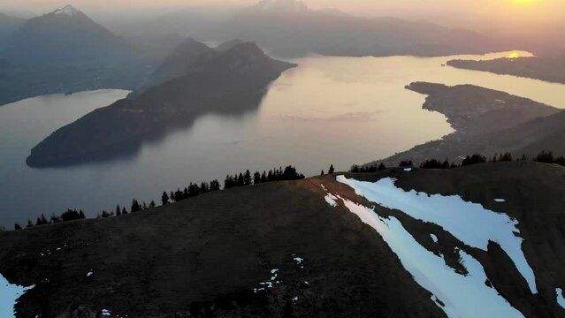 Aerial flyover over the peaks of the Alps near Rigi, Switzerland with a pan up view of Lake Lucerne (Vierwaldstattersee) during a  a golden sunset at the beginning of spring with snow melting
