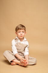 a charming blond four-year-old boy in beige overalls and a white shirt sits on a beige background