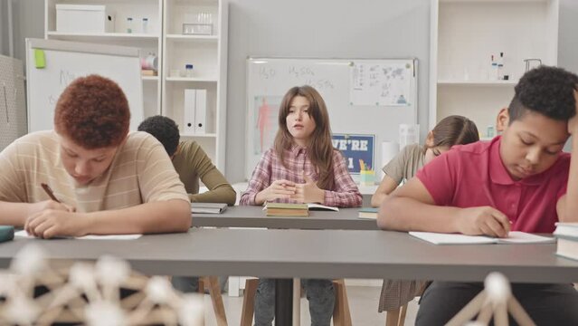 Medium slowmo of Caucasian female pupil raising her hand to ask teacher question sitting at desk with multiethnic classmates during Science lesson