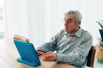 elderly man sitting at table using tablet holding bank card - online banking bills payment