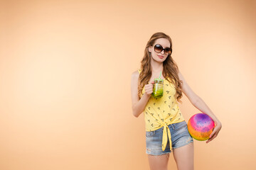 A woman in bright clothes, a yellow T-shirt and denim shorts, sunglasses holds a ball and cold lemonade in her hands, on an isolated background