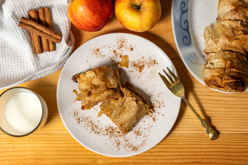 A roll of homemade baked fresh apple pie strudel with an appetizing slice of apple pie on the plate and fresh apples and cinnamon sticks on a wooden table. Rustic breakfast concept. Top View
