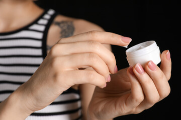 Woman with tattoo holding jar of cream against dark background, closeup