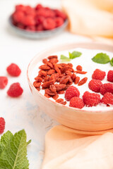 Yogurt with raspberry and goji berries in ceramic bowl on white concrete background. Side view, selective focus.