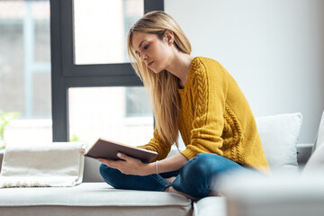 Pretty young woman reading a book while sitting on sofa at home.