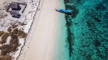 A drone shot of a boat anchored to pink beach on an island near Maumere, Indonesia. There is a fisherman's cottage on the beach, separated by the fence. turquoise coloured water displaying coral reef
