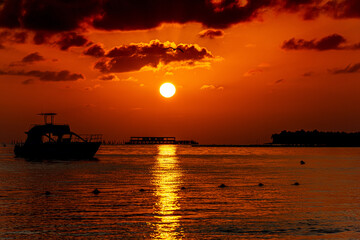 Fototapeta na wymiar Płynący statek niedaleko plaży oraz łódź na morzu podczas wschodu słońca z chmurami i pomarańczowym niebem.