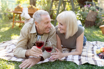 Loving married senior couple drinking wine outside, lying on blanket while having picnic in their garden