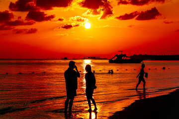 Cień sylwetki pary młodych osób na plaży nad morzem podczas wschodu słońca na pomarańczowym niebie z chmurami, a w tle płynący jacht oraz statek.