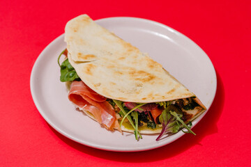 Italian Piadina with prosciutto, sun-dried tomatoes, greens mix, pesto, bechamel and mozzarella. Red background. Close up