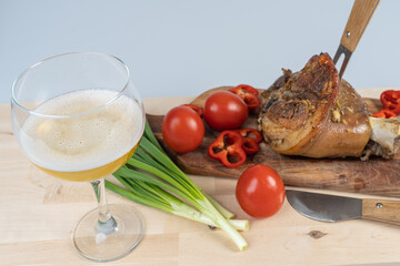 A glass of beer, a fork stuck in a pork knuckle and a knife next to it on a cutting board, meat cooked in the oven with vegetables