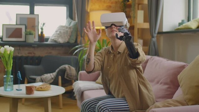 Girl with prosthetic arm sitting on sofa in living room, smiling and touching something in air while experiencing augmented reality with VR headset at home