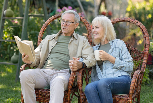Loving elderly couple reading book together, sitting in wicker chairs and spending spring evening in their garden - Powered by Adobe