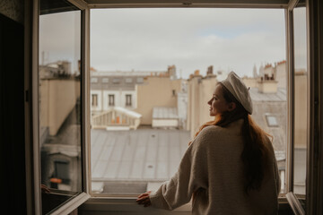 A woman stands near the window against the backdrop of the roofs of Paris. A girl with long hair in a beige suit and beret enjoys the city and the trip