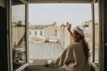 A woman stands near the window against the backdrop of the roofs of Paris. A girl with long hair in a beige suit and a beret enjoys the city and the trip waving to her neighbors