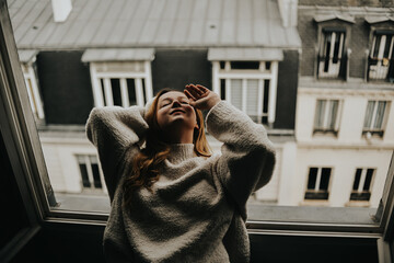 A woman stands near the window against the backdrop of the roofs of Paris. Girl with long hair in a beige suit posing