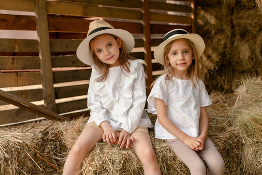 Two smiling little girls sitting on haystack in hayloft on summer day