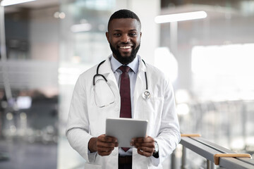 Positive young black doctor holding digital tablet, clinic interior