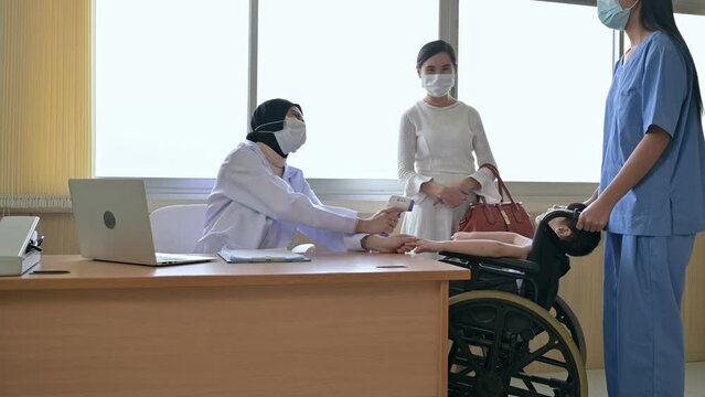 Asian female pediatrician wearing face mask using forehead thermometer checking sick girl sitting on wheelchair with mother and nurse assitant in clinic