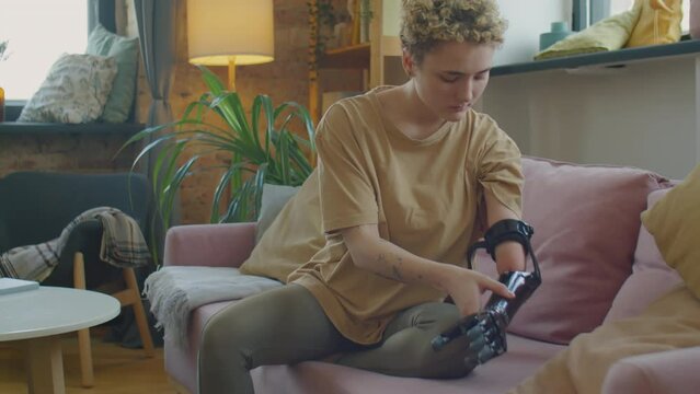 Tilt up shot of Caucasian girl sitting on sofa at home and putting on prosthetic arm