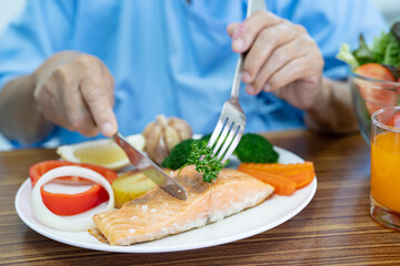 Asian senior or elderly old lady woman patient eating Salmon steak breakfast with vegetable healthy food while sitting and hungry on bed in hospital.