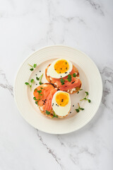 Sandwich with delicious toppings, smoked salmon, eggs, herbs and microgreens radish, black sesame seeds over white plate on white marble table background. Healthy open sandwich superfood. Top view.