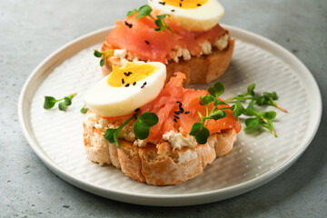 Sandwich with delicious toppings smoked salmon, eggs, herbs and microgreens radish, black sesame seeds over white plate on gray concrete table background. Healthy open sandwich superfood. Top view.
