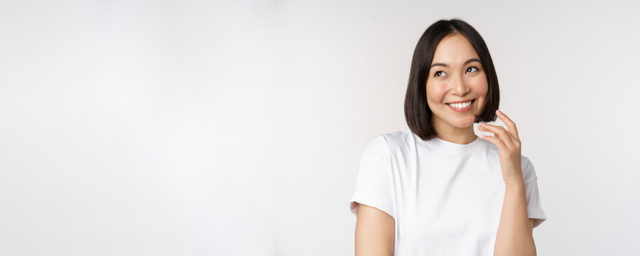 Portrait of cute coquettish woman laughing and smiling, looking aside thoughtful, thinking or imaging smth, standing in white t-shirt over studio background