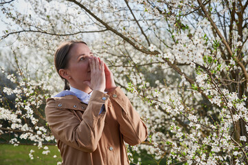 Woman allergic suffering from seasonal allergy at spring, posing in blossoming garden at springtime. Young woman sneezing in front of blooming tree. Spring allergy concept