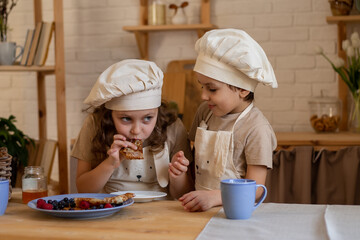 A boy and a blonde girl at the table in aprons and chef's hats are eating pancakes