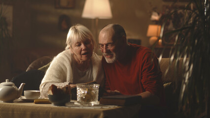 Happy aged husband and wife smiling and discussing photos in album while sitting on couch near table in morning in cozy living room at home