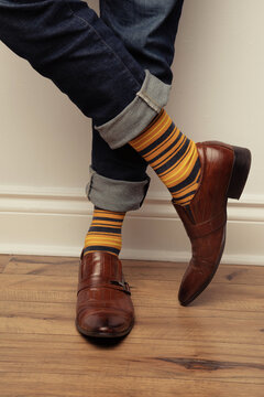 Studio photo of a pair of men's feet wearing a pair of yellow and brown striped socks, brown leather shoes, and jeans that are rolled up. 